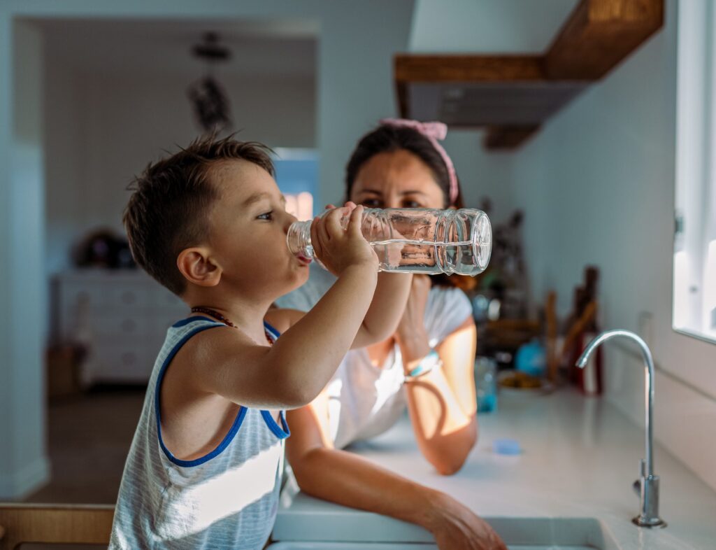 A child drinks from a bottle of water in his kitchen, next to his mom.