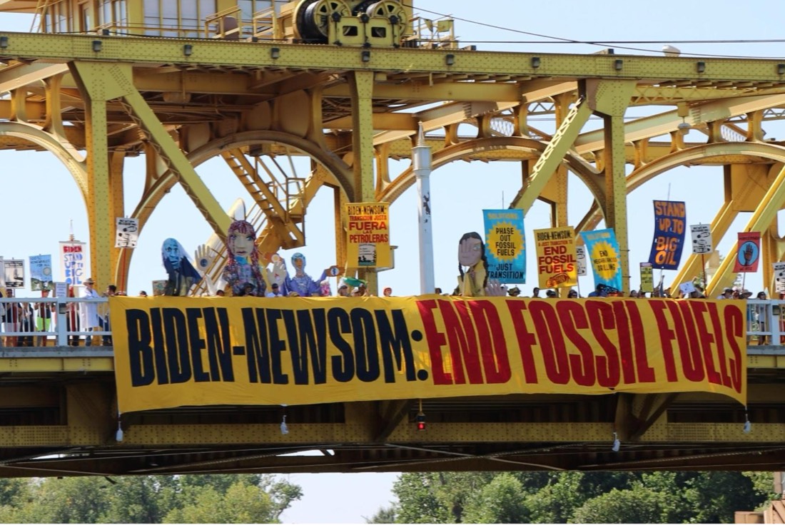 Protesters on a bridge holding up signs and a banner that reads "Biden Newsom End Fossil Fuels"