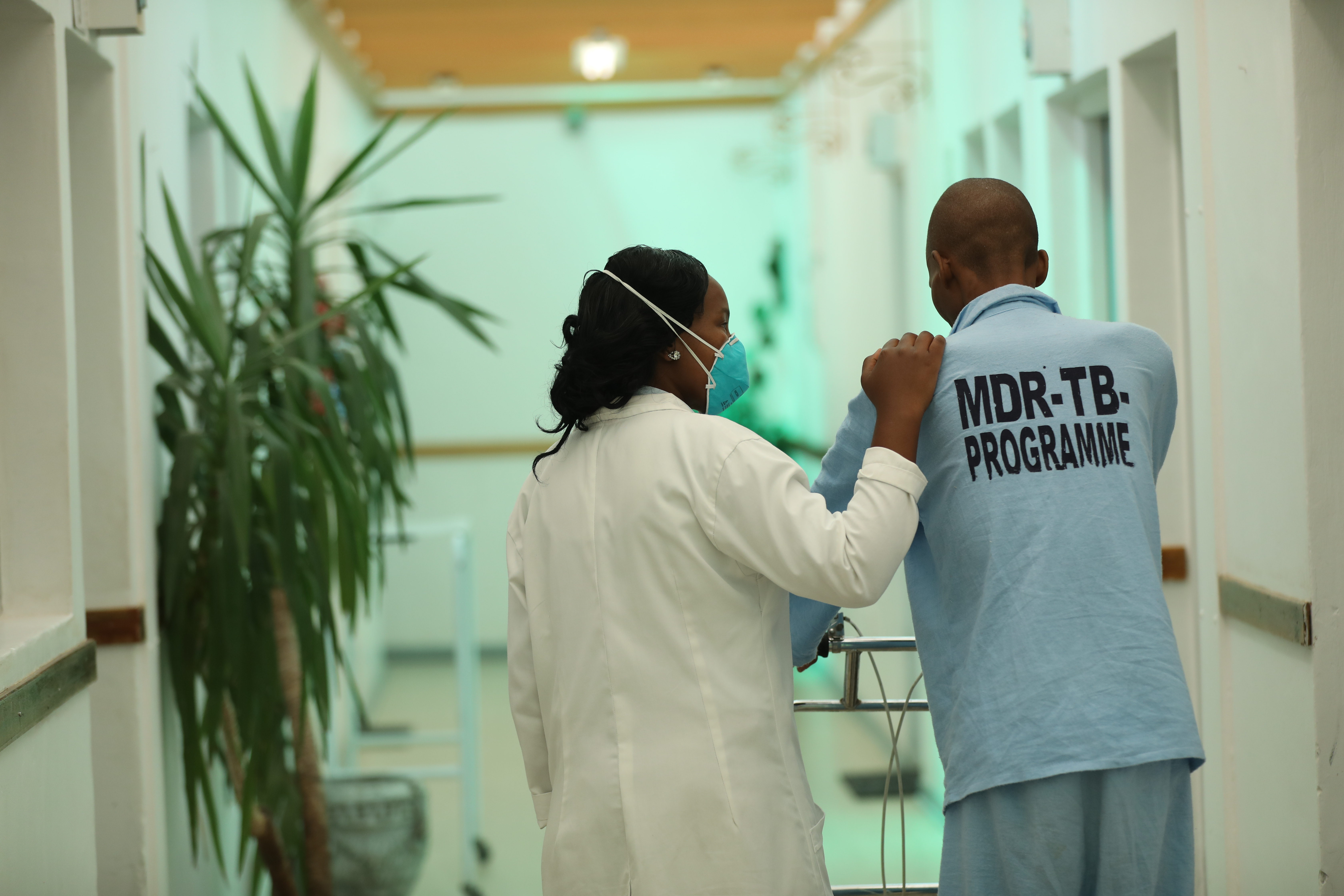 A masked doctor accompanying a tuberculosis patient down a hospital hall in Lesotho.